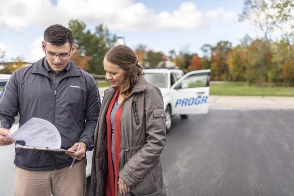 Recruiting: Man and woman review paperwork in front of Progressive vehicle