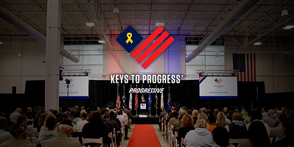 The Keys to Progress and Progressive logos above a person standing at a podium in front of a seated audience.