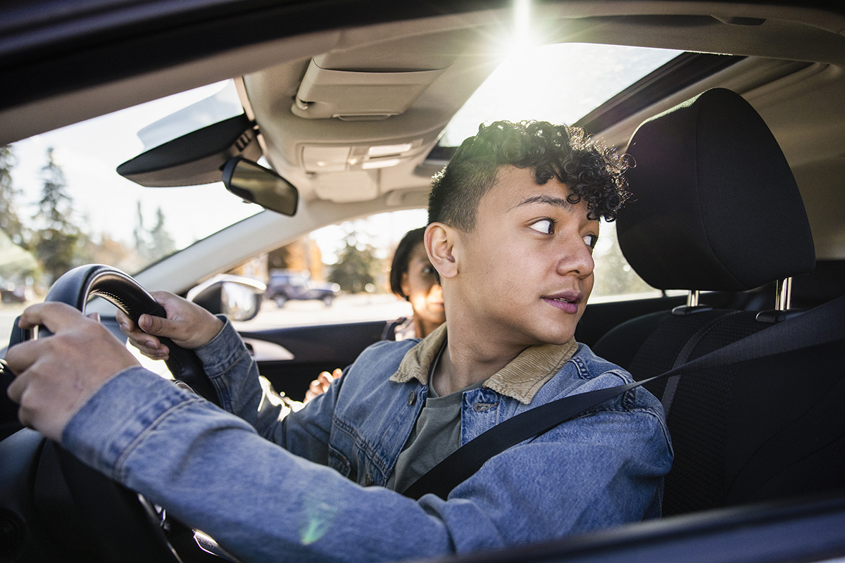 A teen driver wearing a seatbelt in the driver's seat of a car looks over his left shoulder out the back window.