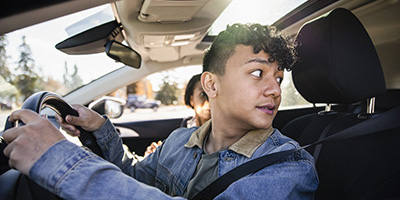 A teen driver wearing a seatbelt in the driver's seat of a car looks over his left shoulder out the back window.