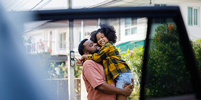 A man holding a little girl, both smiling, standing on the other side of an open car window in a neighborhood.