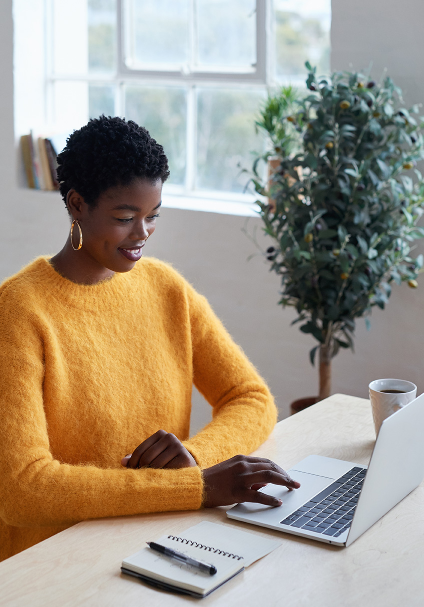 A smiling woman sits at a desk with a laptop, a notebook and pen, and a mug.