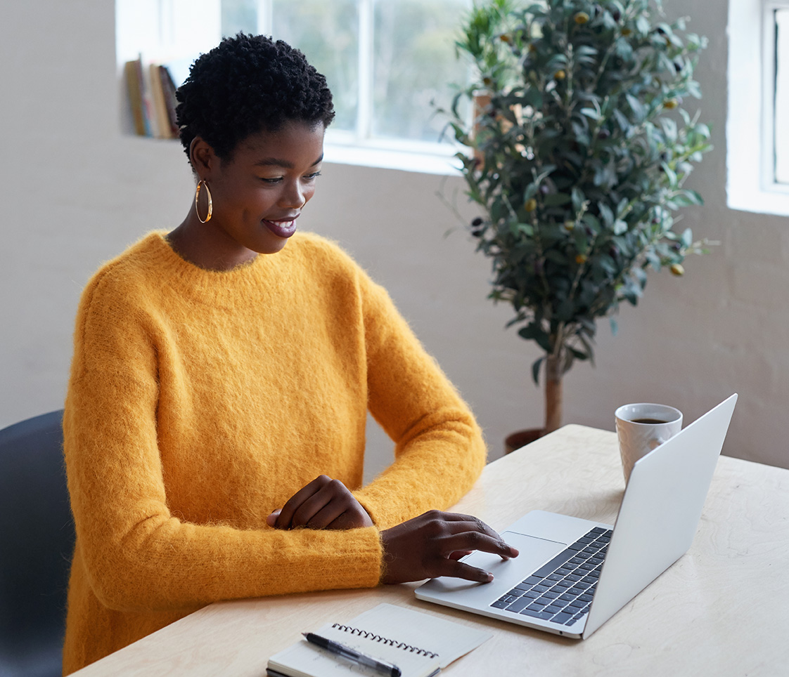 A smiling woman sits at a desk with a laptop, a notebook and pen, and a mug.