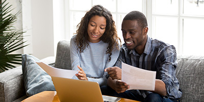 A couple sitting on a couch hold papers and gesture toward a laptop sitting in front of them.